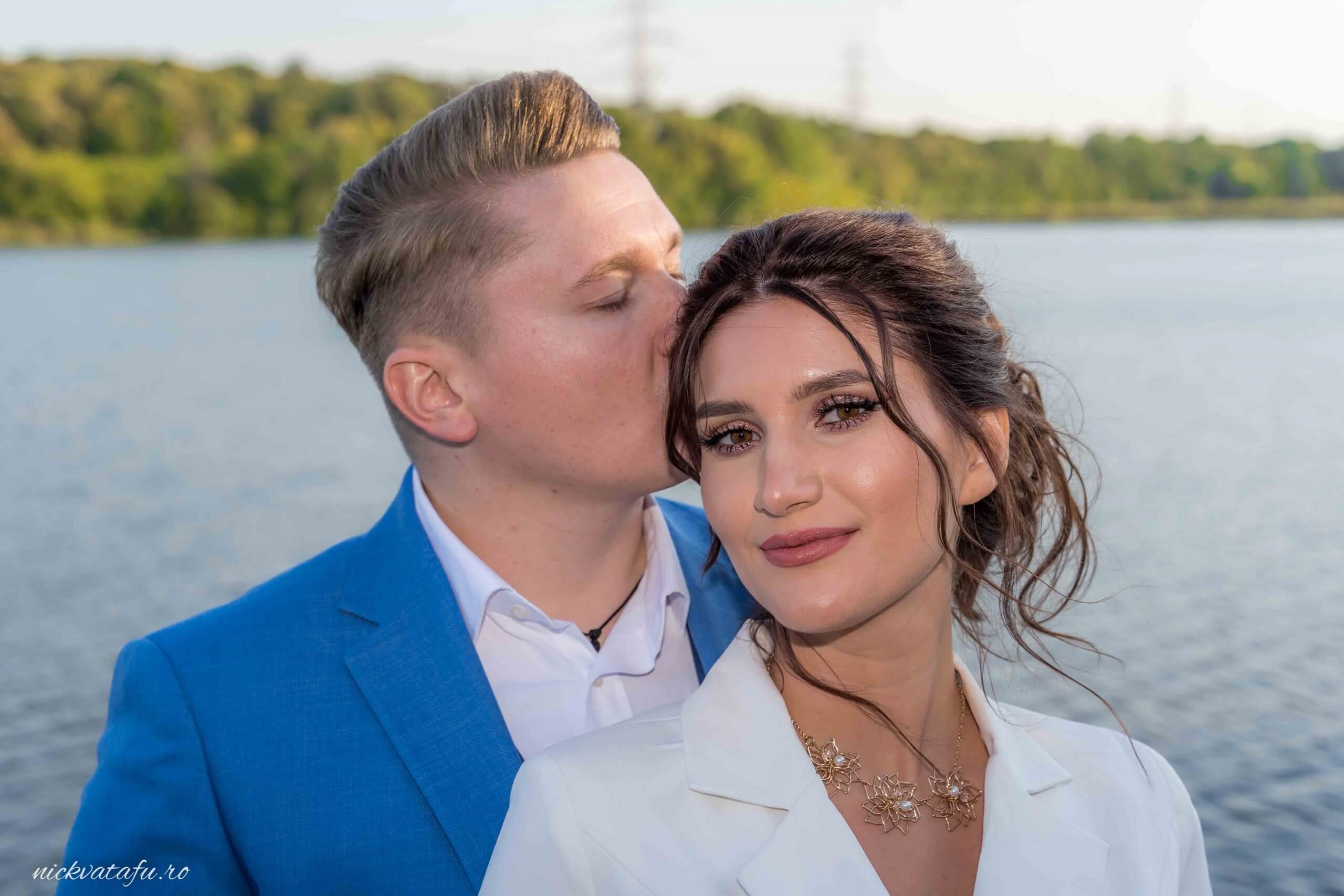 Mire și mireasă zâmbind, cu un sărut delicat pe frunte, fotografie de nuntă la malul apei, plină de emoție.