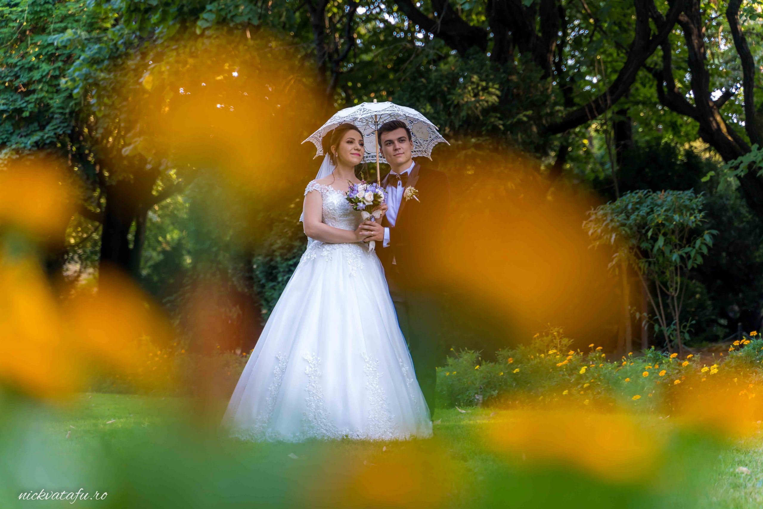 Mire și mireasă zâmbind sub un umbrelă elegantă, înconjurați de flori, fotografie de nuntă în natură.