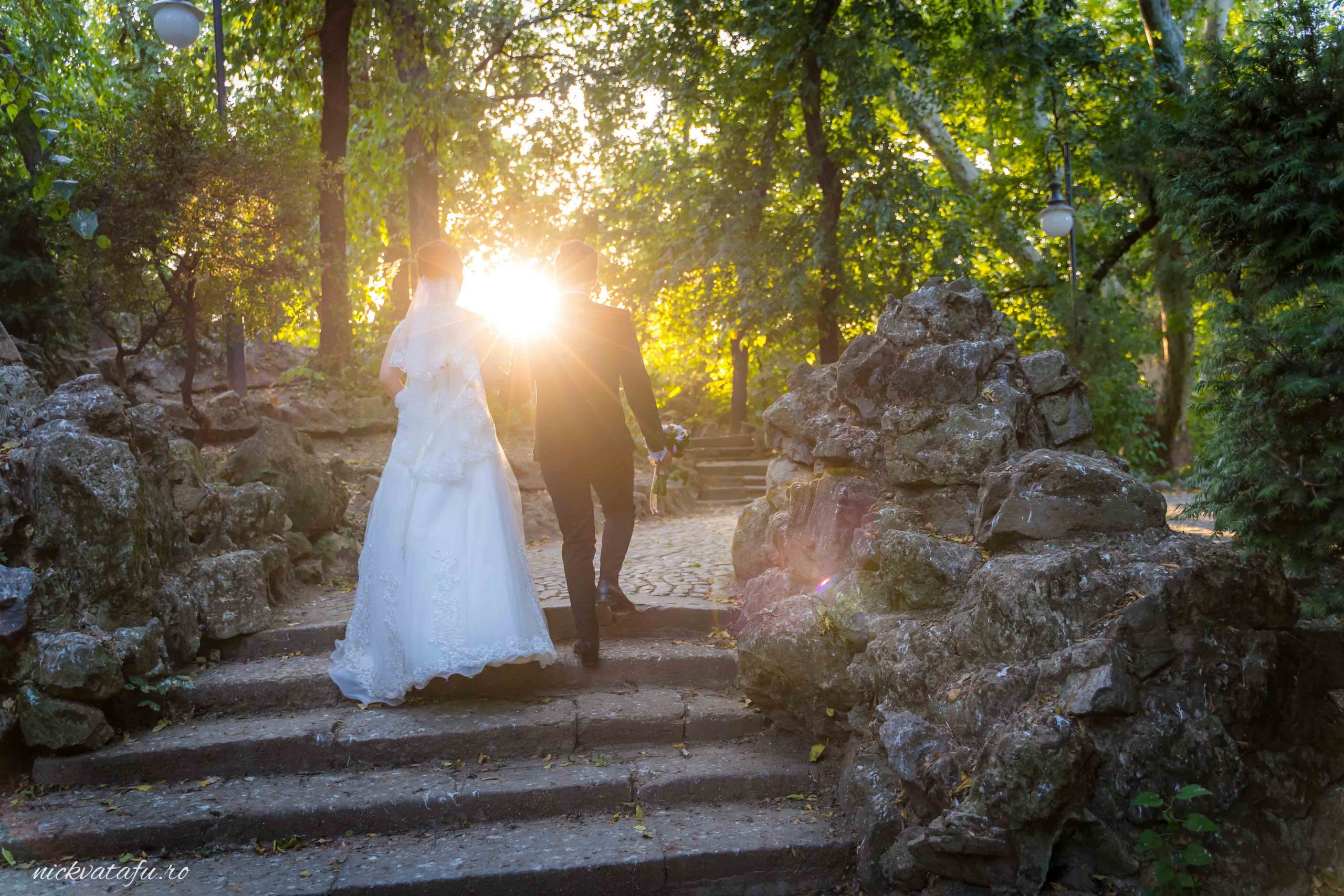 Mire și mireasă urcând pe scări într-o atmosferă romantică, cu soarele strălucind printre copaci, fotografie de nuntă.