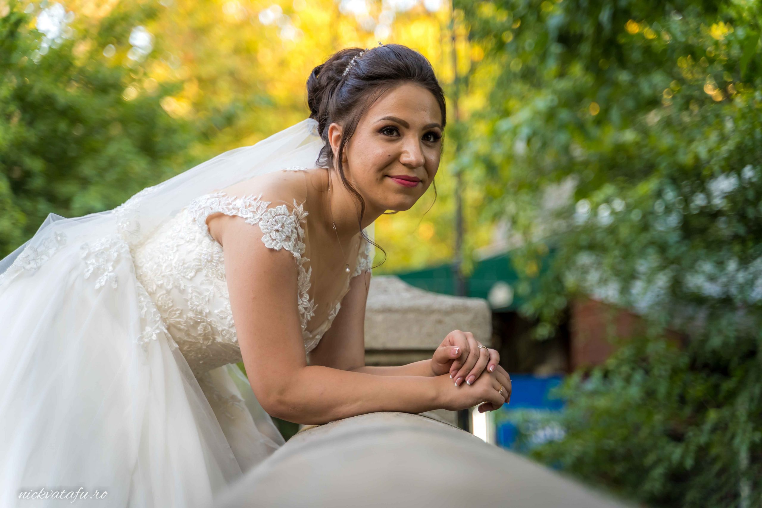 Mireasă zâmbitoare, așezată pe o balustradă, înconjurată de natură, fotografie de nuntă plină de emoție.