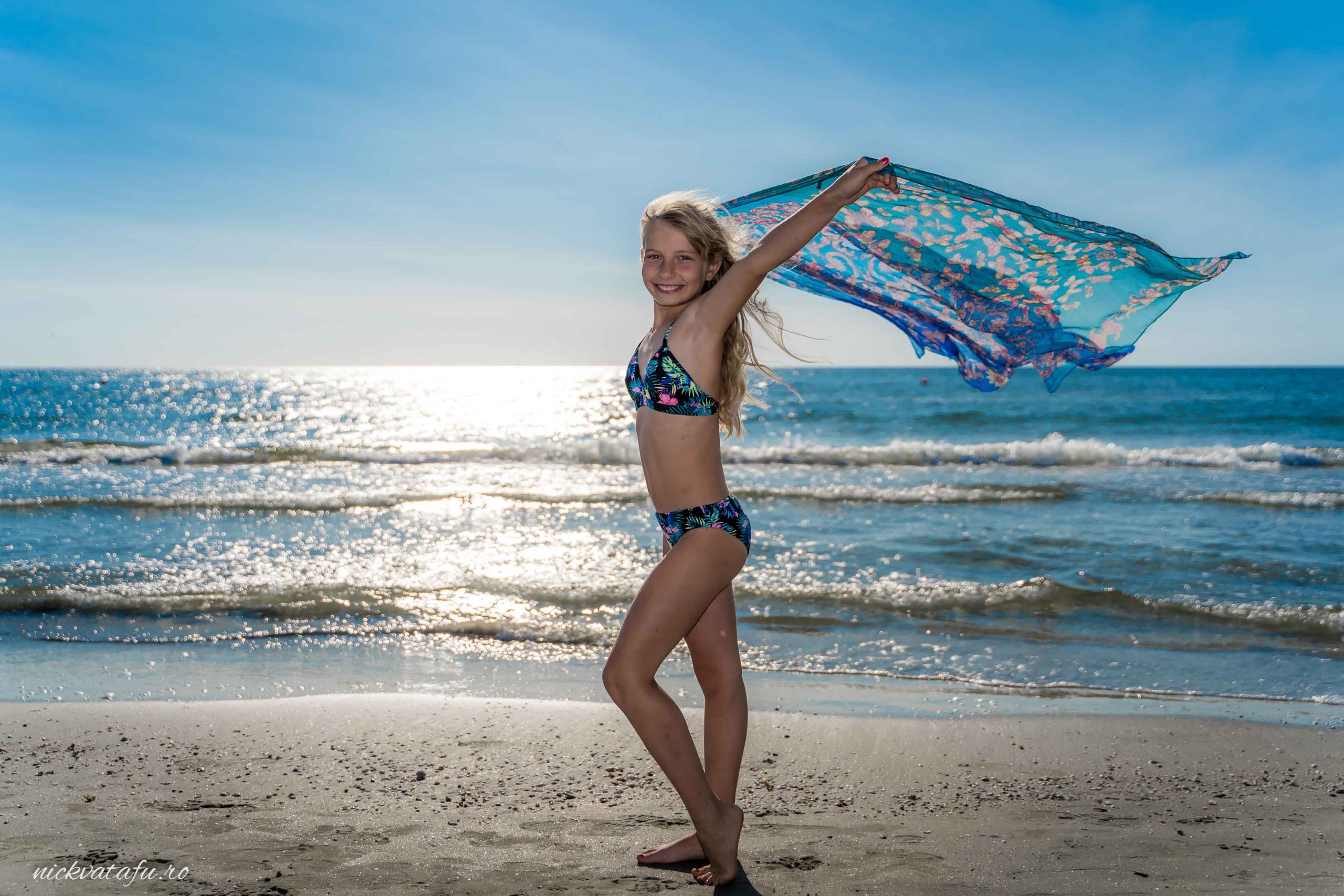 Fată jucându-se pe plajă, cu un șal colorat în vânt, evocând bucurie și libertate.