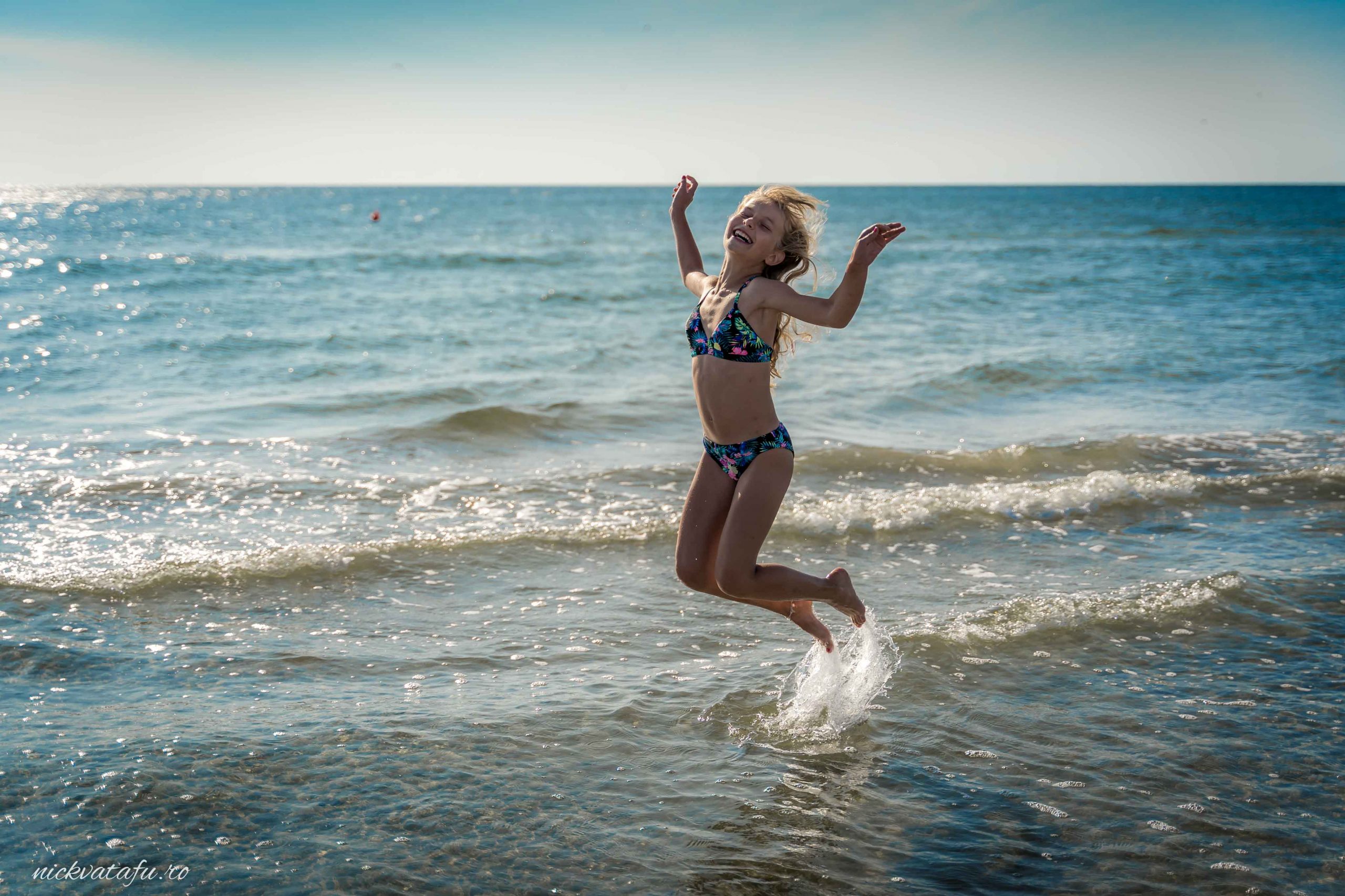 Fată zâmbitoare sărind în apă, emanând bucurie și energie, pe o plajă însorită.