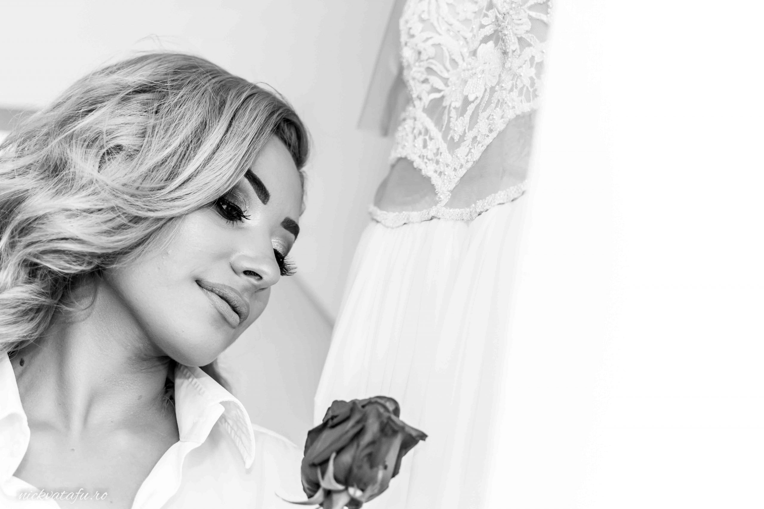 Mireasă contemplând o floare roșie, cu rochia de nuntă în fundal, fotografie elegantă și emoționantă.