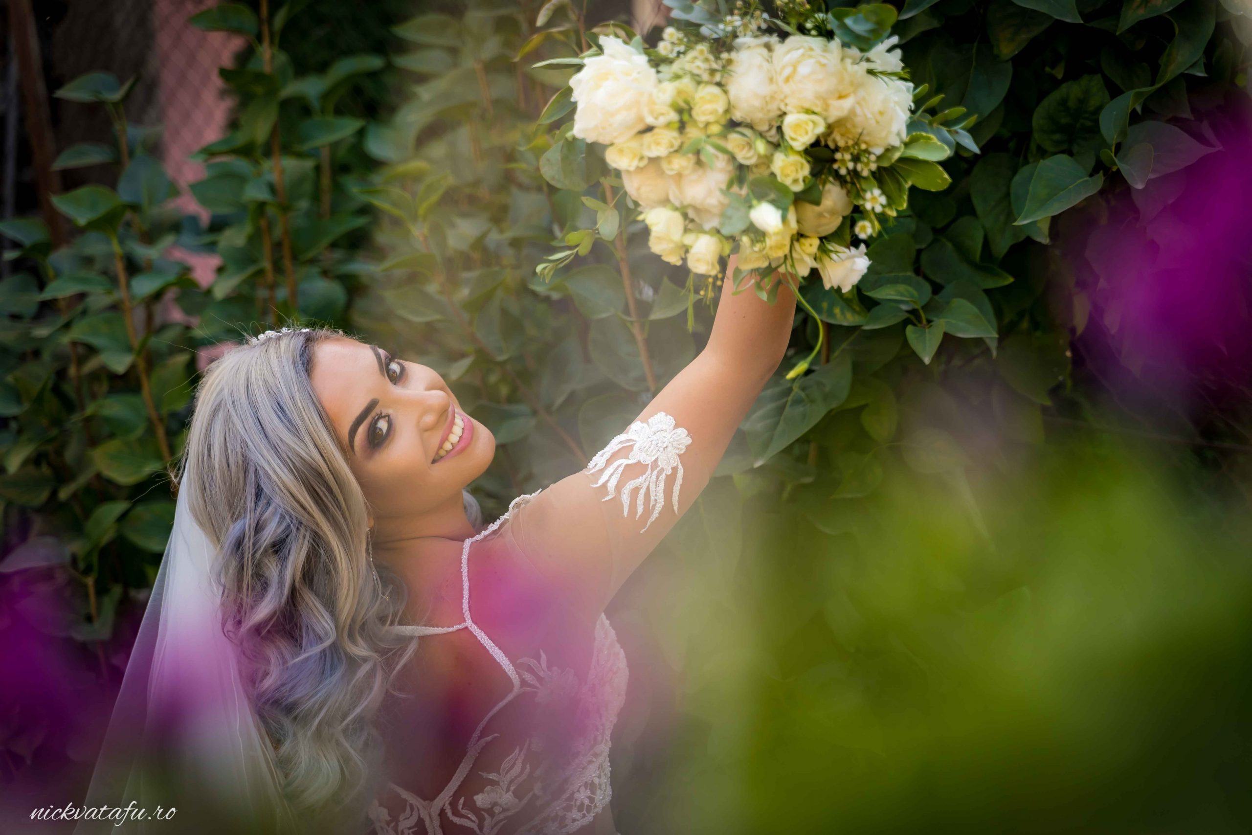 Mireasă zâmbitoare, cu un buchet de flori, într-un decor natural plin de verdeață, fotografie de nuntă elegantă.