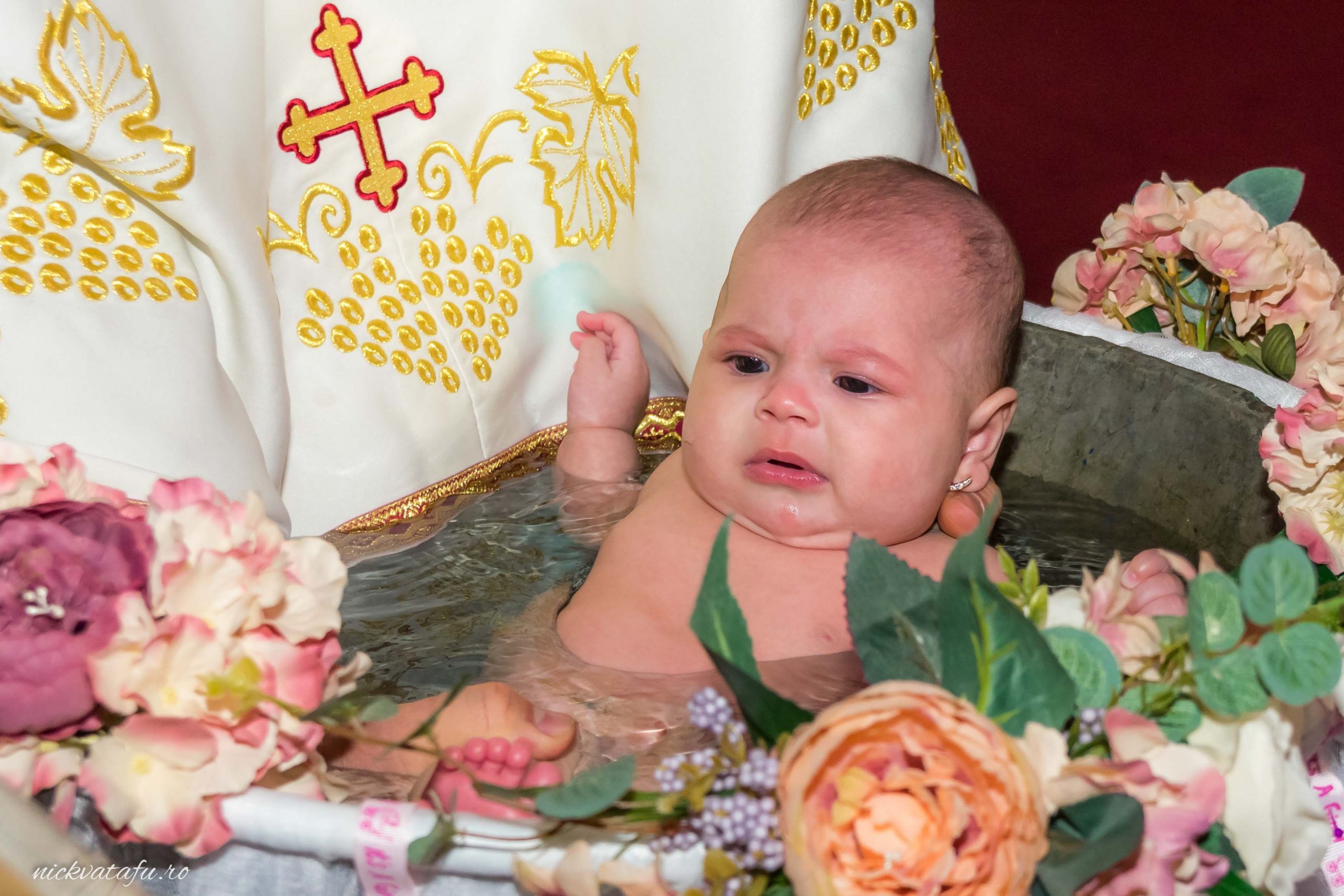 Bebeluș în apă, în momentul botezului, înconjurat de flori, fotografie de botez Pitești plină de emoție.