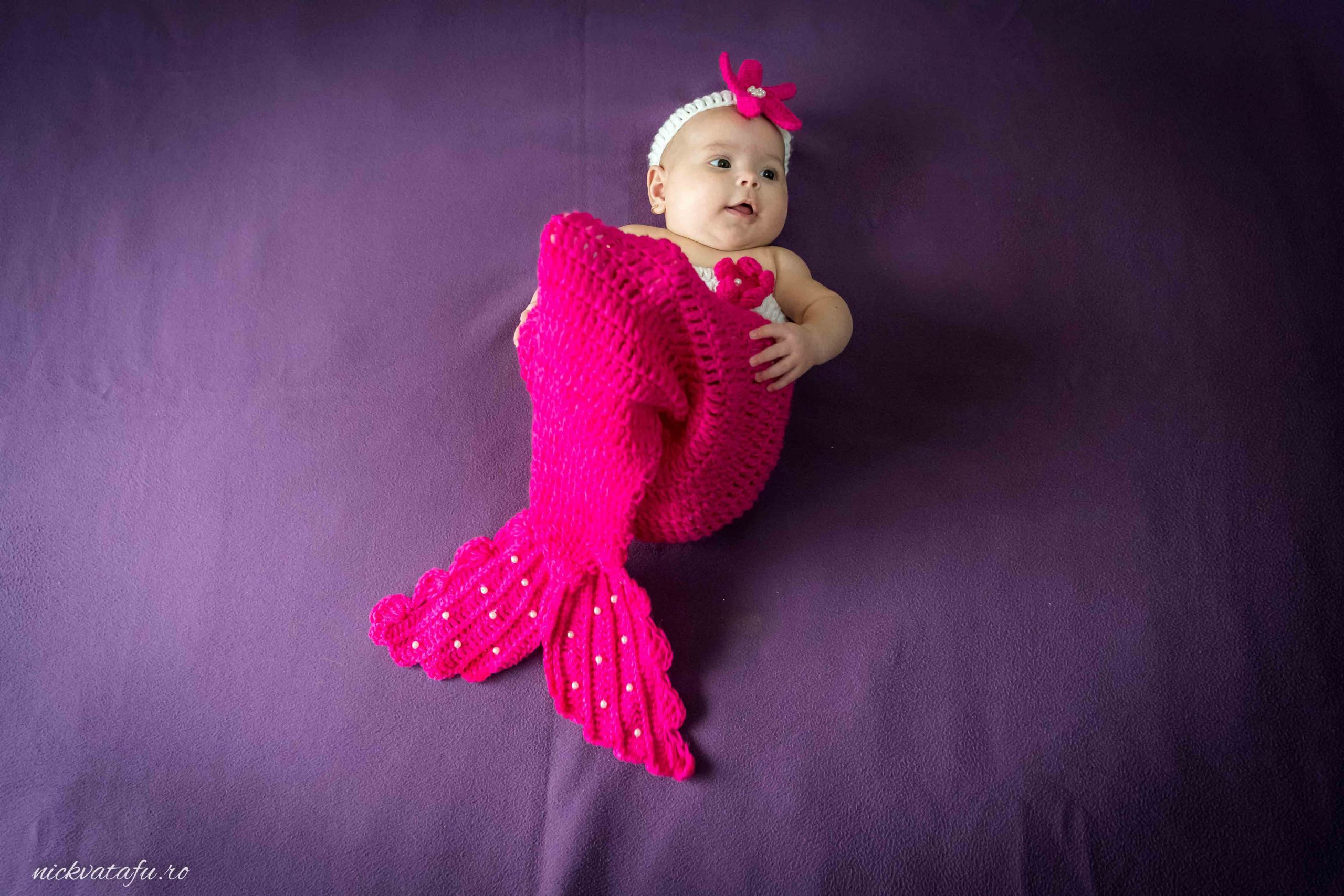 Bebeluș îmbrăcat în costum de sirenă roz, pe un fundal violet, radiază bucurie și inocență.