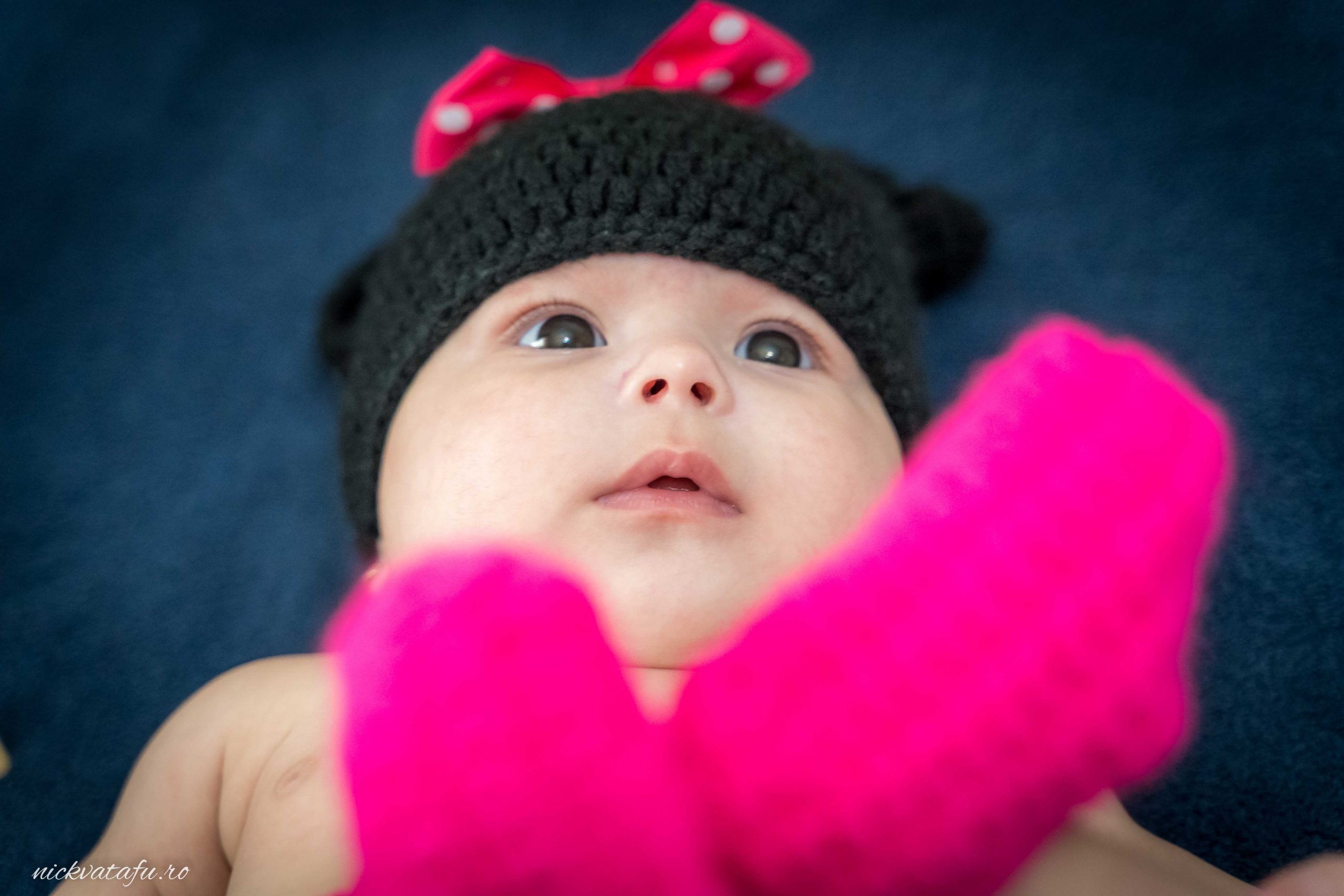 Bebe cu pălărie neagră și fundiță roșie, privind cu curiozitate, fotografie de botez plină de emoție.