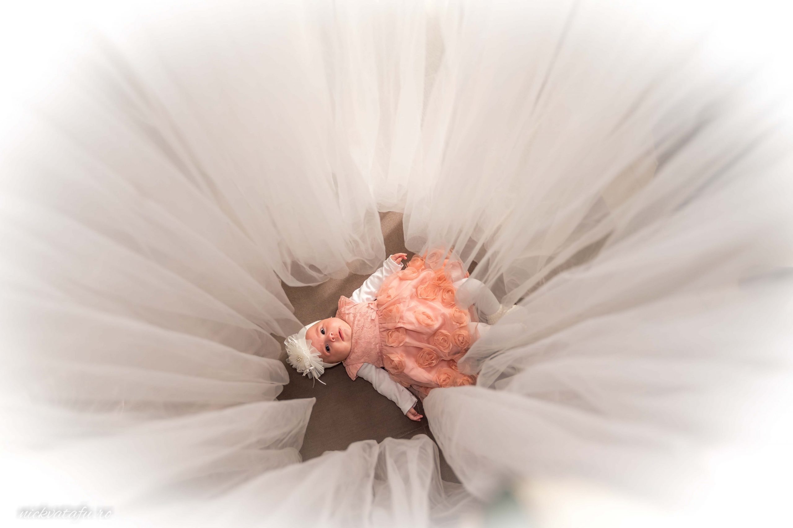 Fetiță în rochie de botez, zâmbind și jucându-se în mijlocul unui voal alb, fotografie de botez emoționantă.