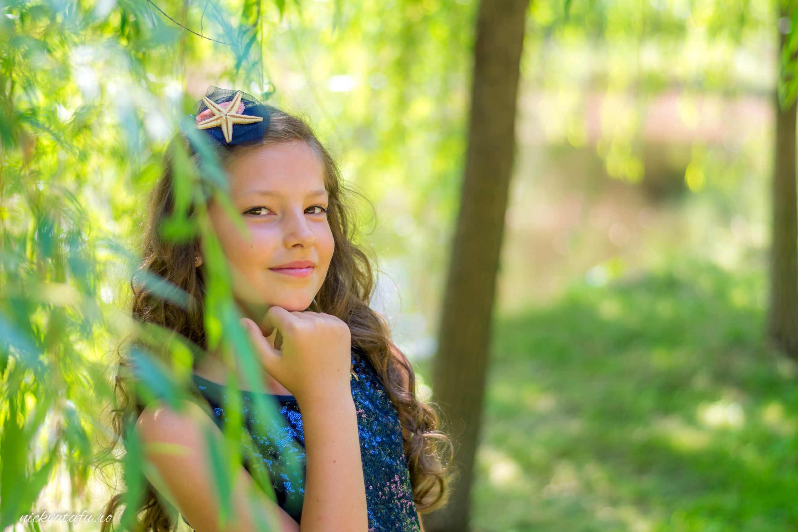 Fetiță cu zâmbet jucăuș, pozând printre frunzele verzi, capturând emoția unui botez în natură.