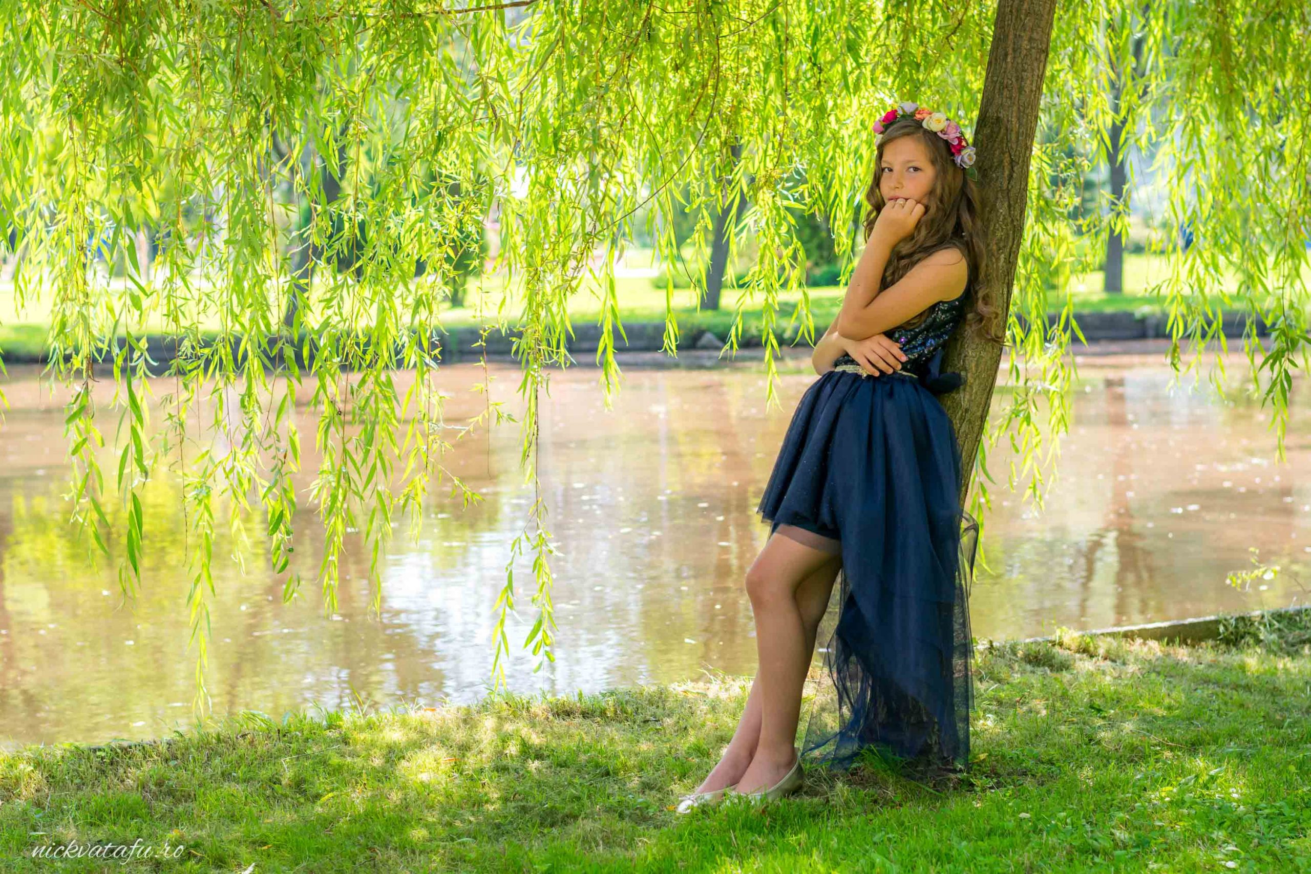 Fată elegantă, cu rochie albastră, stând lângă un copac, pe malul unui lac, emanând emoție și naturalețe.