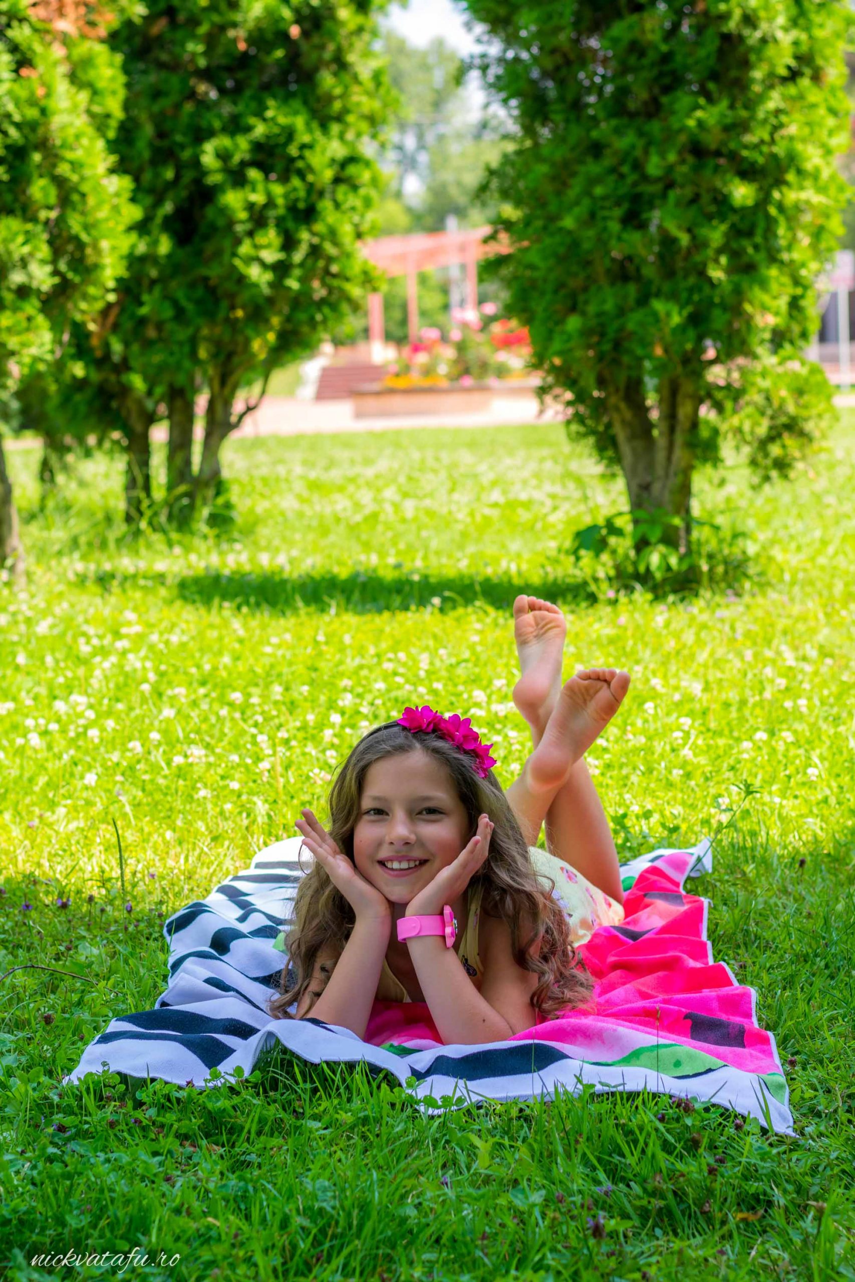 Fetiță zâmbitoare cu flori în păr, relaxată pe o pătură în parc, emanând bucurie și inocență.