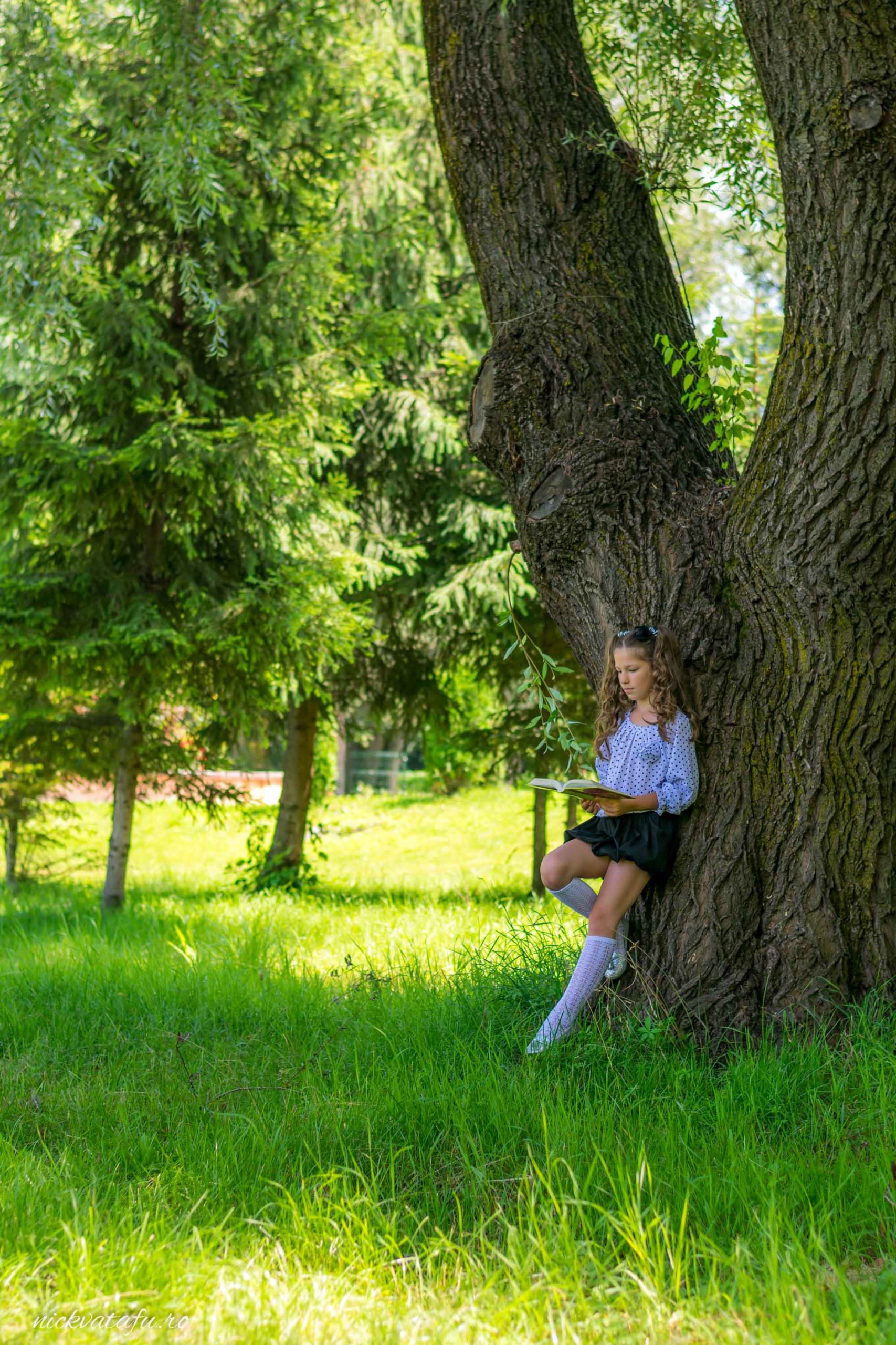 Fată zâmbitoare, relaxată, stând lângă un copac mare într-un peisaj verde, capturând o atmosferă de bucurie și liniște.