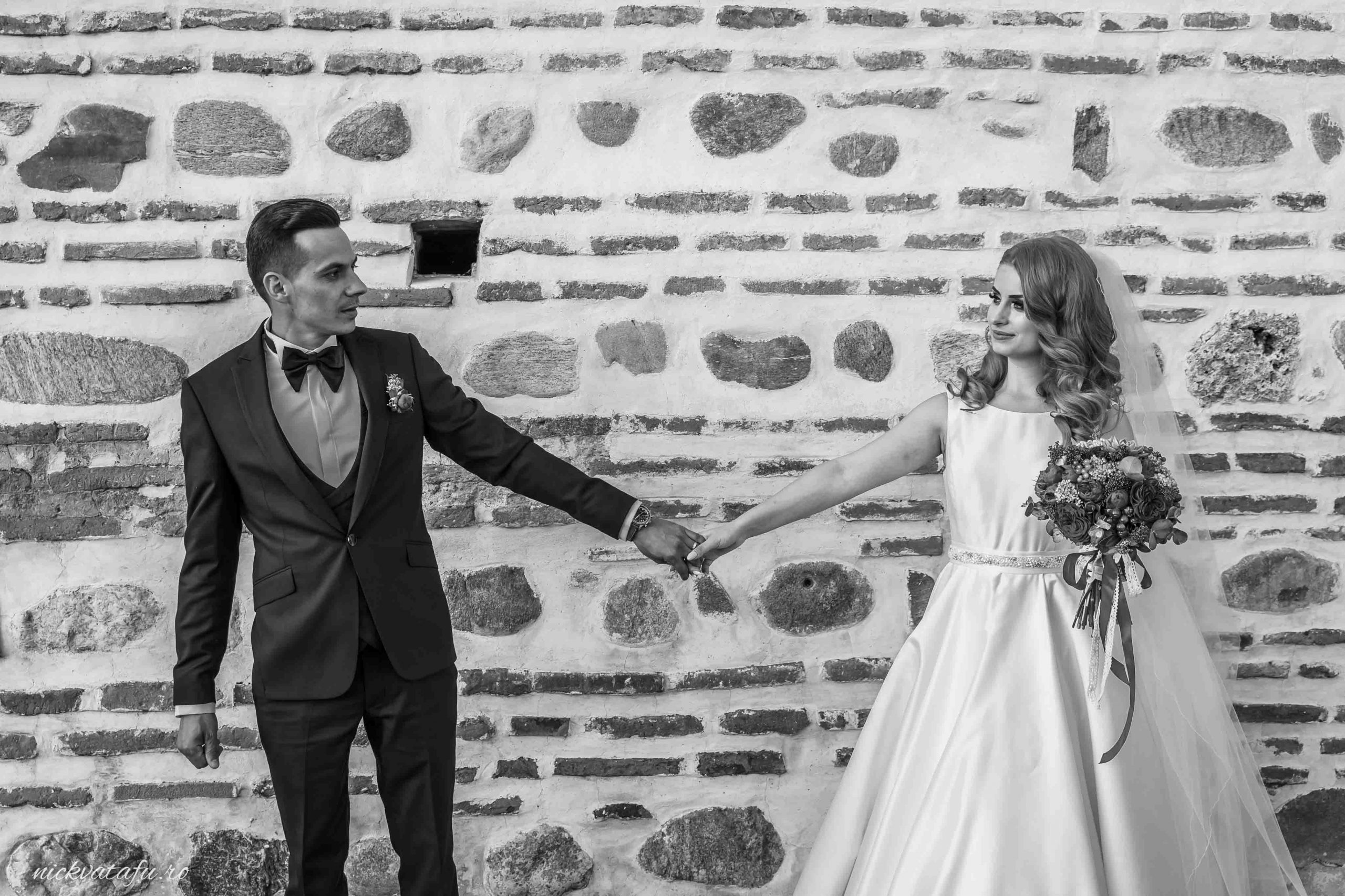 Mire și mireasă ținându-se de mână, zâmbind emoționați, în fața unei pereți cu piatră, fotografie de nuntă elegantă.