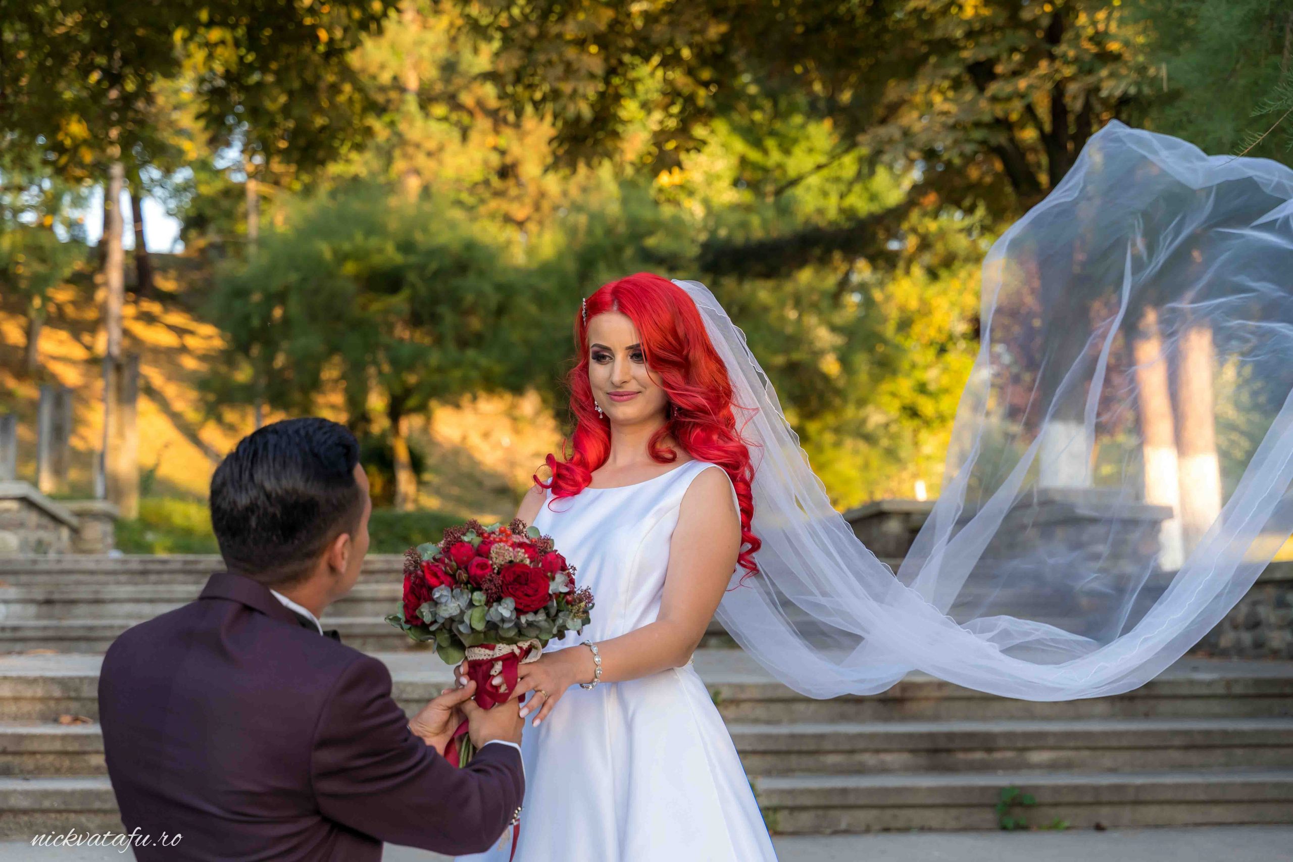 Mireasă cu păr roșu vibrant și buchet de flori, primind o cerere în căsătorie într-un cadru natural, fotografie de nuntă plină de emoție.