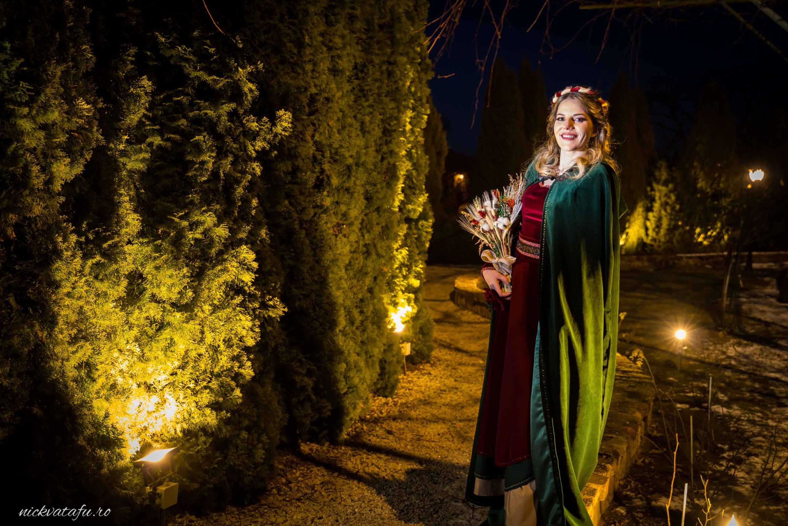 Fată elegantă, îmbrăcată în rochie de gală, ținând un buchet, sub lumini calde într-o grădină bine întreținută.