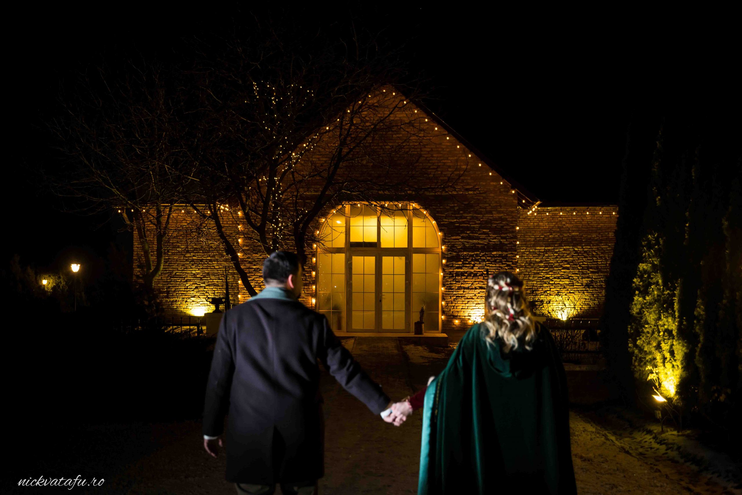 Mire și mireasă ținându-se de mână, îndreptându-se spre o locație elegant iluminată, în atmosferă romantică de nuntă.