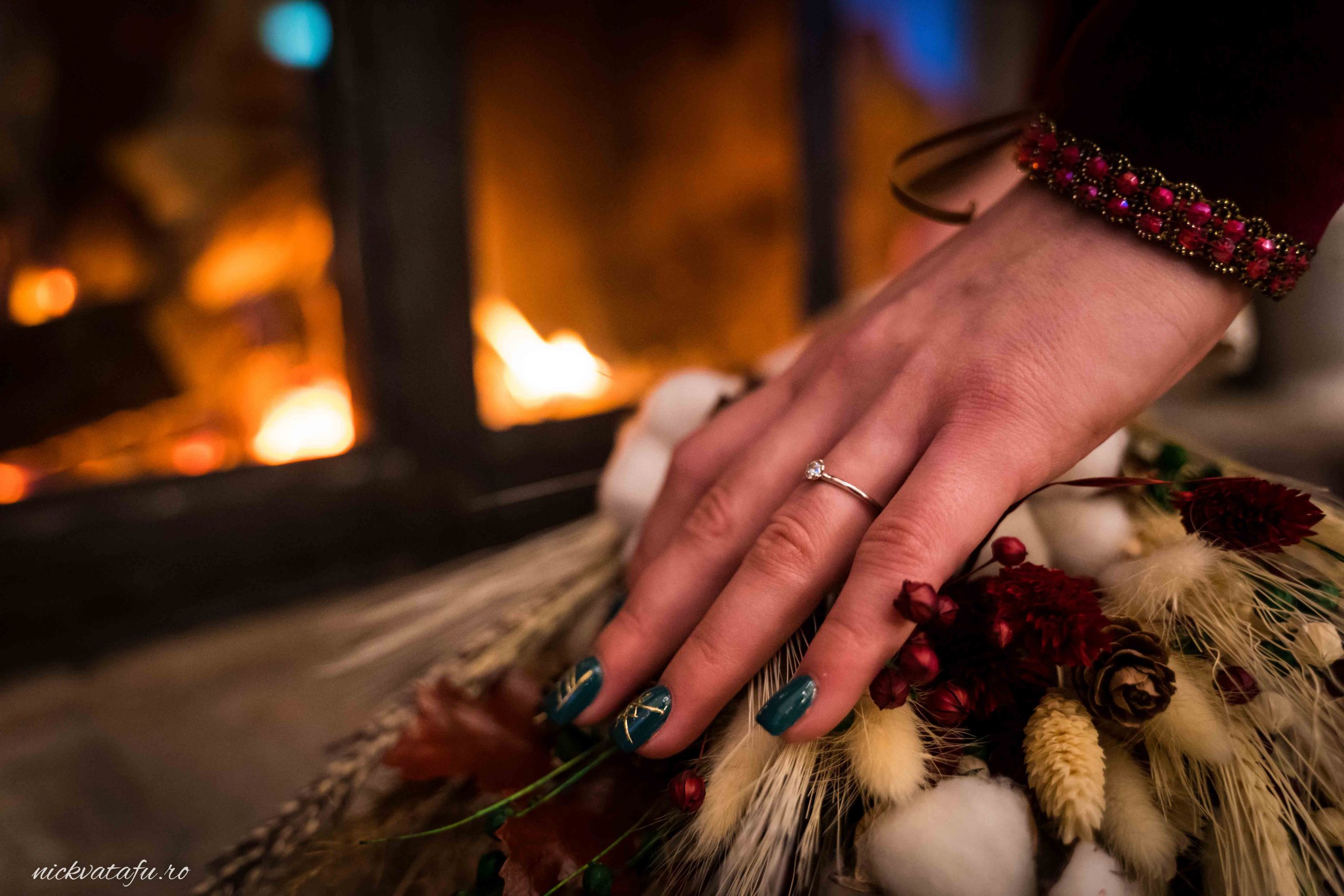 Mână cu inel, așezată delicat pe un buchet de flori, în fundal un foc cald, detalii nuntă elegante.