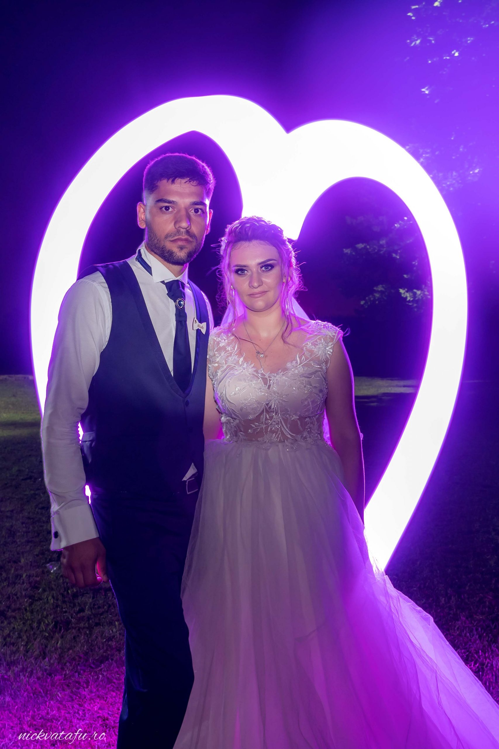 Mire și mireasă zâmbind fericiți în fața unui decor luminos în formă de inimă, fotografie nuntă plină de emoție.