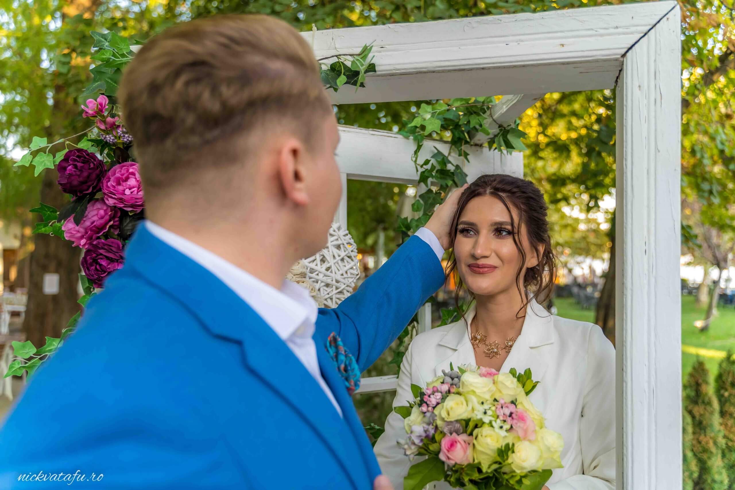 Mire și mireasă într-un moment de tandrețe, cu flori în mână, la o ceremonie de nuntă în aer liber.