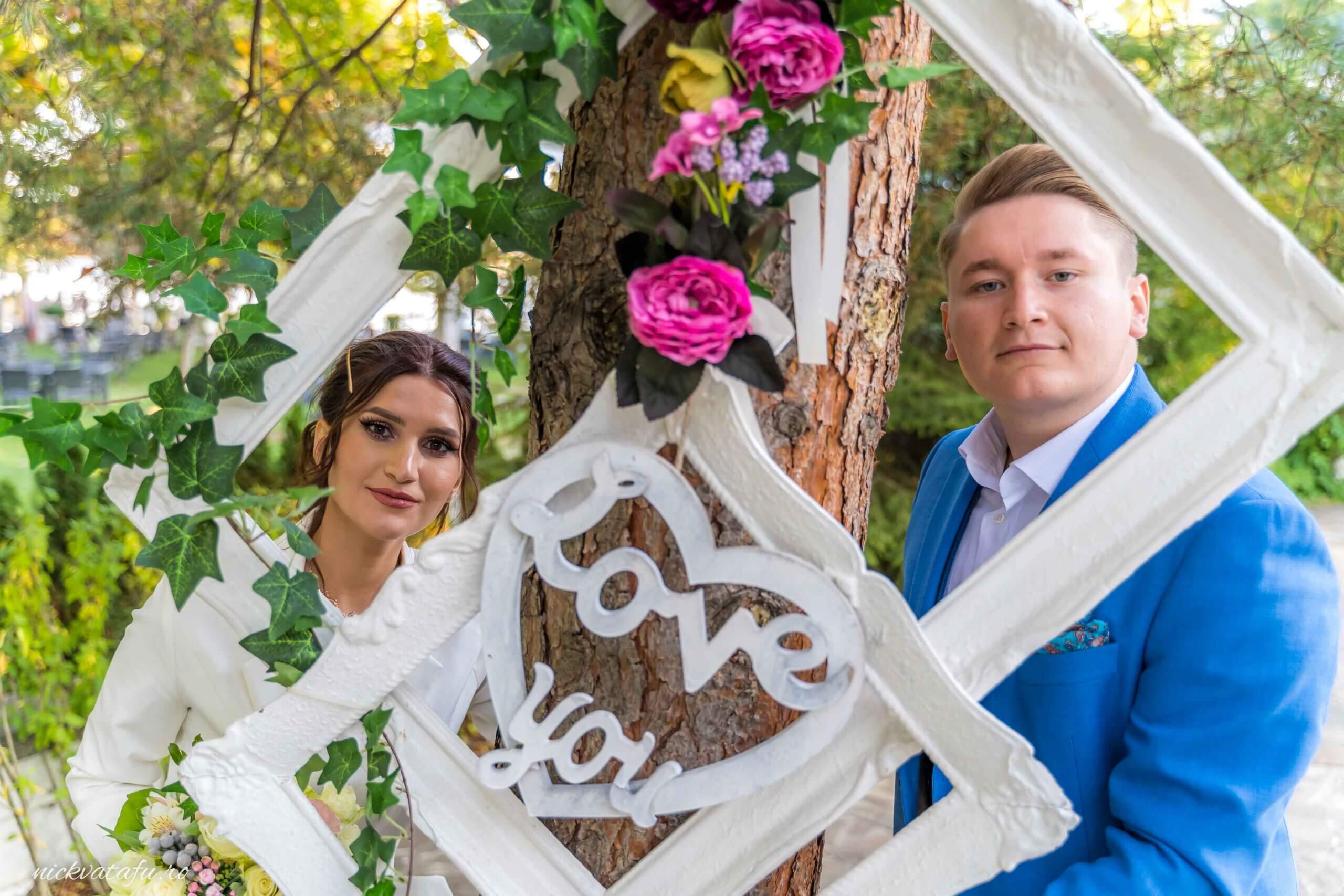 Mire și mireasă zâmbind fericiți în fața unui decor floral, fotografie de nuntă plină de emoție și iubire.